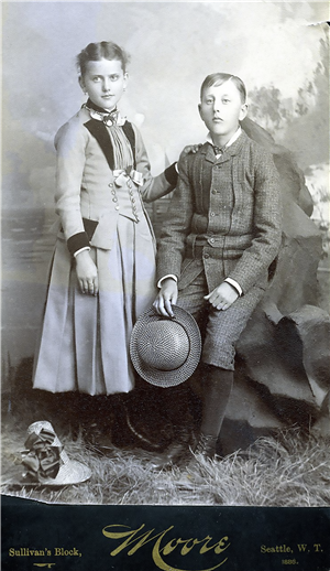 Two young adults one standing in a dress and one seated in wool coat and knickers pose in front of a large rock on a floor of straw